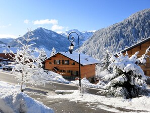 A wooden chalet with a sloped roof, surrounded by snow-covered trees and mountains in the background.