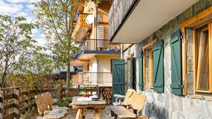 Una terraza de madera con un muro de piedra, mobiliario exterior y un balcón con contraventanas verdes.