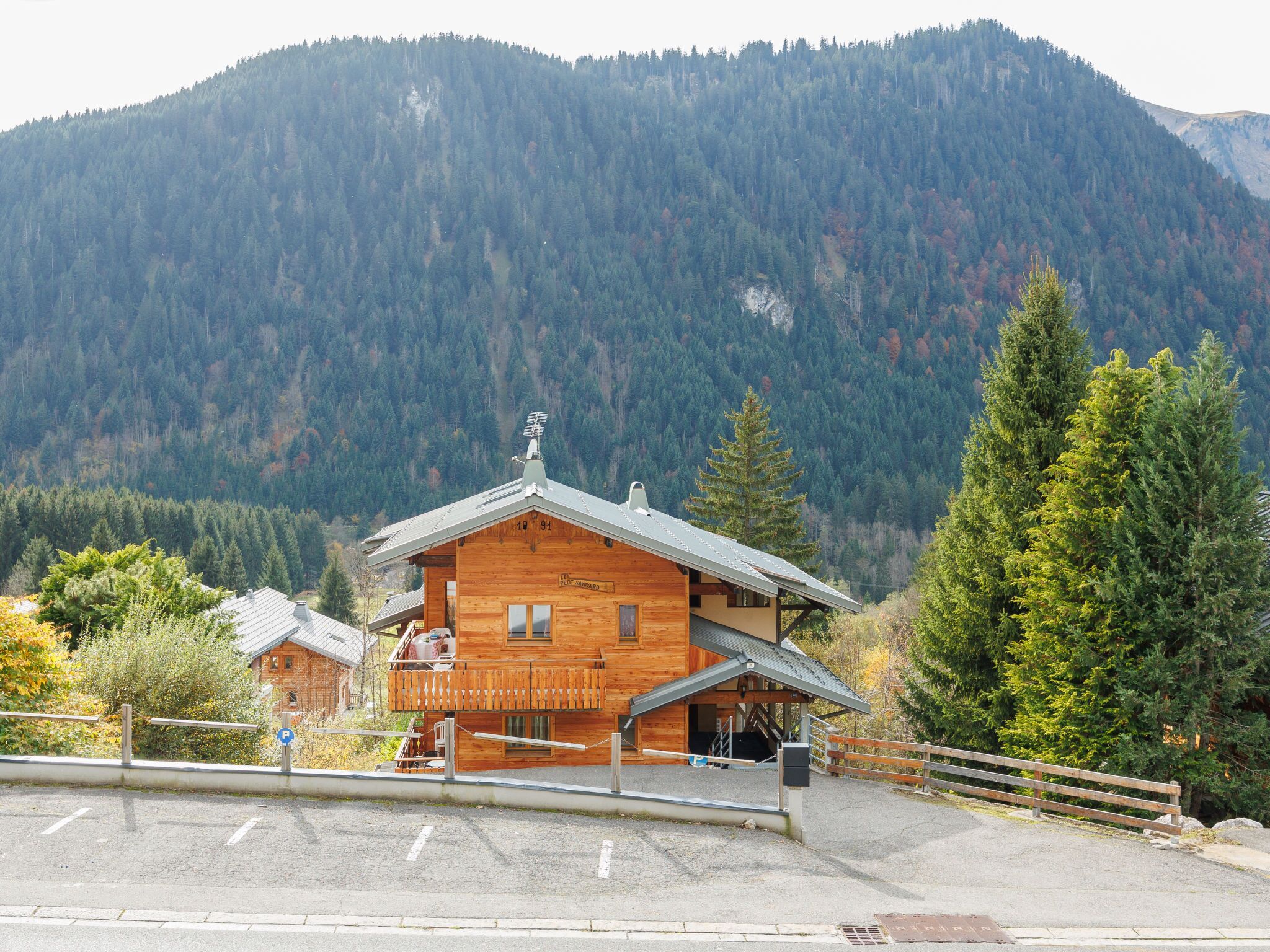 A wooden lodge with a balcony, surrounded by a parking area and a mountain backdrop.