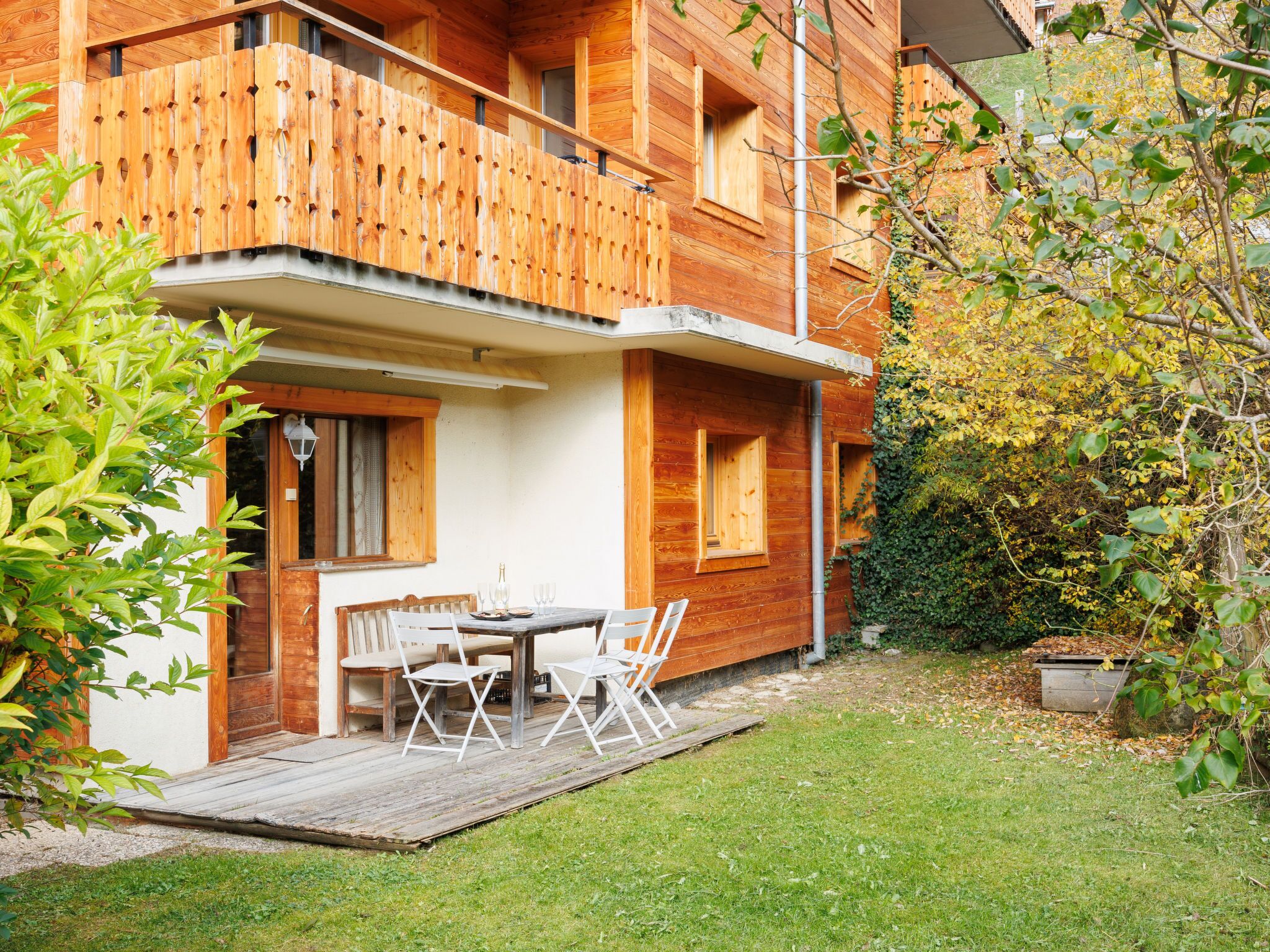 A wooden cabin with a balcony, a dining area with chairs and a table, and a garden with a wooden deck.