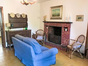 A living room with a brick fireplace, a blue sofa, wicker chairs, a wooden cabinet, and wall-mounted artwork.
