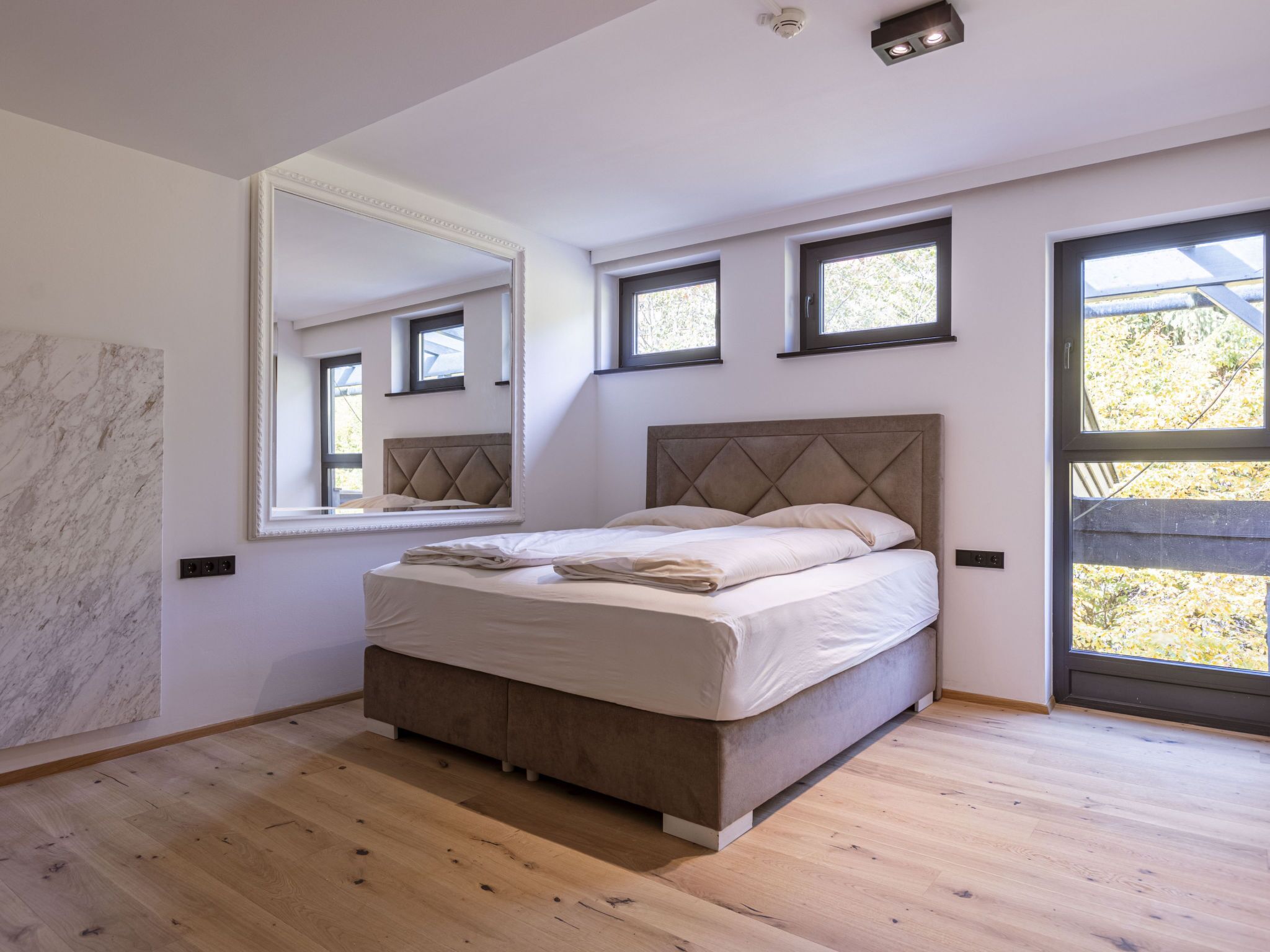 A modern bedroom with a large bed, a mirror, and multiple windows.