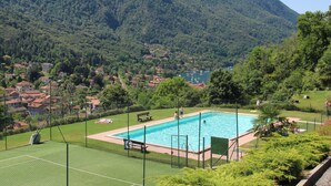 Une piscine entourée d’un espace délimité par une clôture, avec un court de tennis, le tout situé dans un cadre de paysages montagneux.