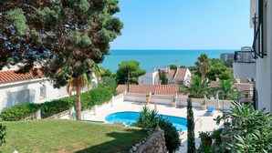 A pool area with a view of the sea, surrounded by greenery and buildings with red-tiled roofs.