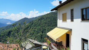 Ein weißes Gebäude mit gelbem Vordach, einem Balkon mit Blumentöpfen und Blick auf eine bewaldete Bergkette.