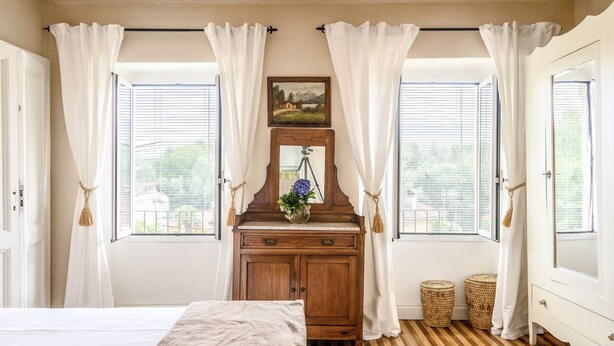 A bedroom with a wooden dresser, a bed with a brown blanket, and two windows with white curtains. - Rachele by Interhome (Verbania)