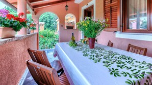 A covered outdoor dining area with a table set for two, wooden chairs, potted plants, and a view of greenery through an archway.