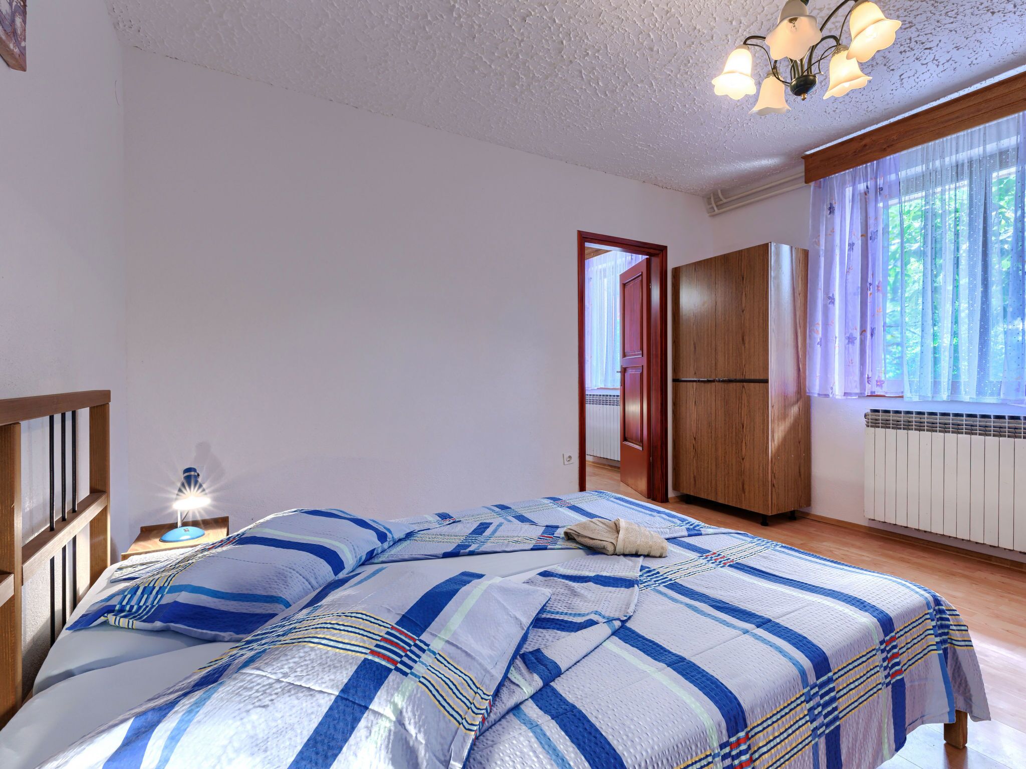 A bedroom with a bed, a nightstand, a wooden wardrobe, and a window with curtains.