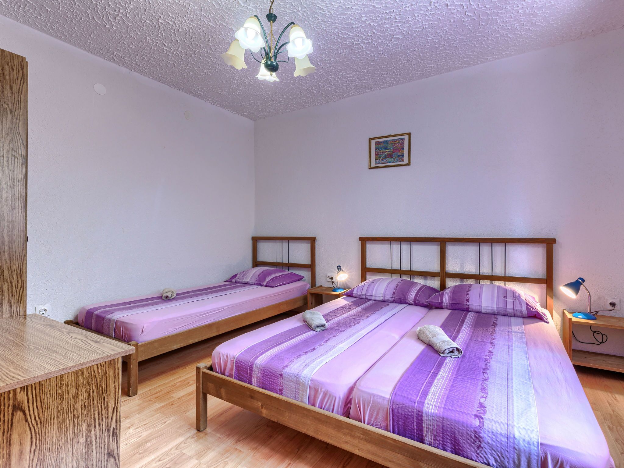 Ein Zimmer mit zwei Einzelbetten, einem Holztisch, einem kleinen Tischchen, einem Wandbild und einer Deckenleuchte.