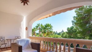 A balcony with wicker furniture, a glass table, and a view of palm trees and greenery.