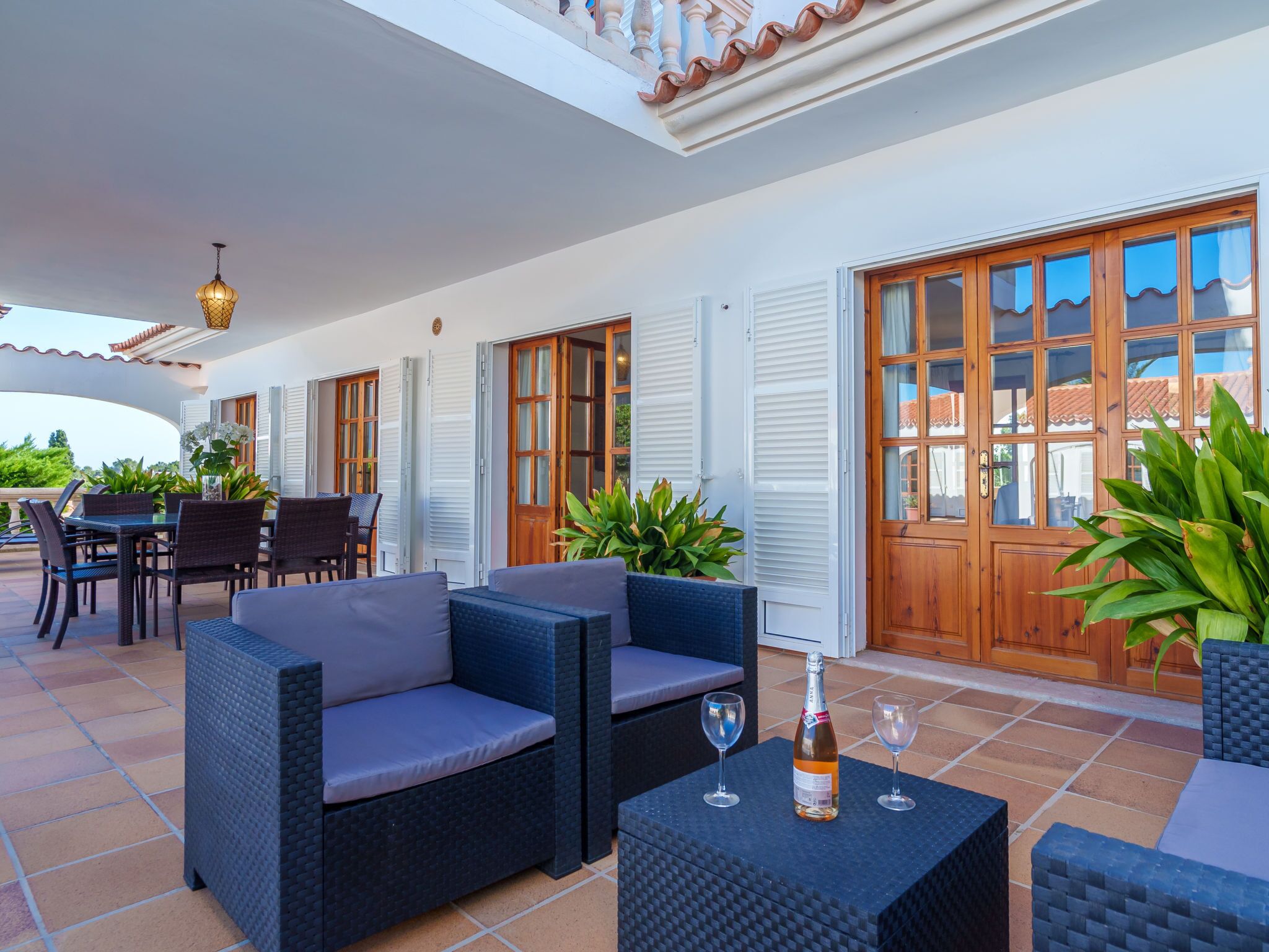 A covered outdoor patio with wicker furniture, a table with a bottle of wine, and a view of the house through large windows.
