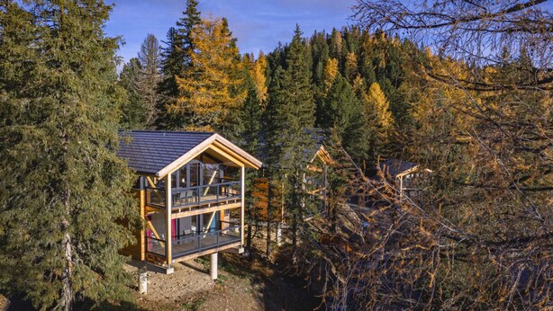 A cabin with a wooden exterior, large windows, and a balcony, surrounded by dense forest and autumn foliage.