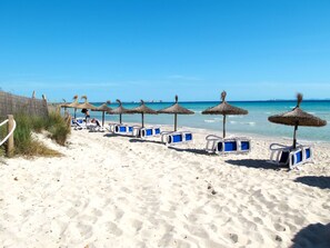 Ein Strand mit Reihen von Liegestühlen und Reetsonnenschirmen, ein sandiger Uferstreifen und ein klares, blaues Meer.