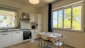 Una cocina moderna con armarios blancos, una mesa de comedor para cuatro personas y grandes ventanales con vistas a árboles.