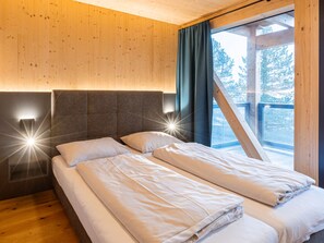 A double bed with a headboard and bedside lamps, a window with curtains, and a view of trees outside.
