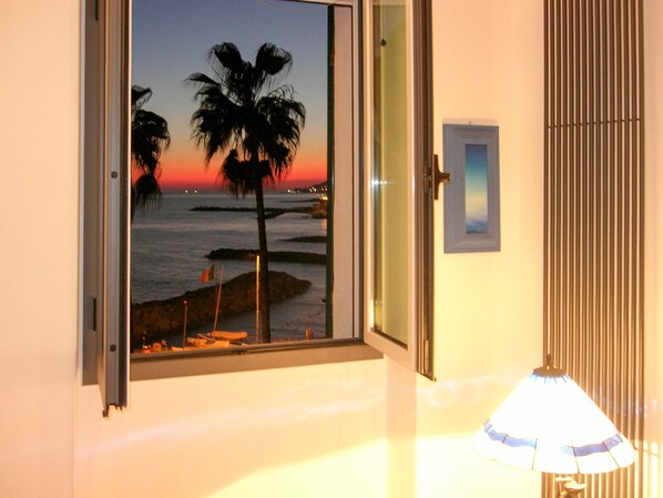A room with a large window overlooking a beach at sunset, a lamp, and a door.