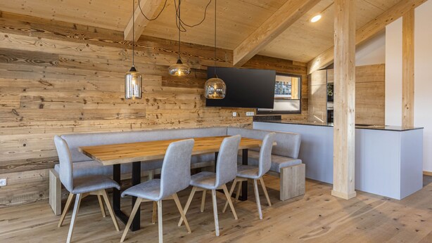 A modern kitchen with a wooden wall, a dining area with a wooden table and grey chairs, and a built-in island. - 02_NTH005_Sup Chalet mit Sauna und Hot Tub 2 Etage by Interhome (Turracher Höhe)