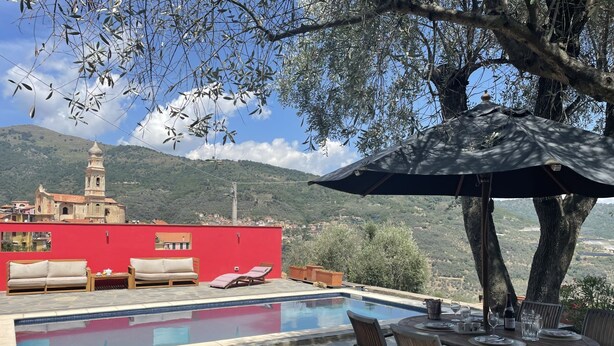 A pool area with lounge chairs and a table, overlooking a red building and a hill with a tower. - Il Rifugio (BCM110) by Interhome (Boscomare)