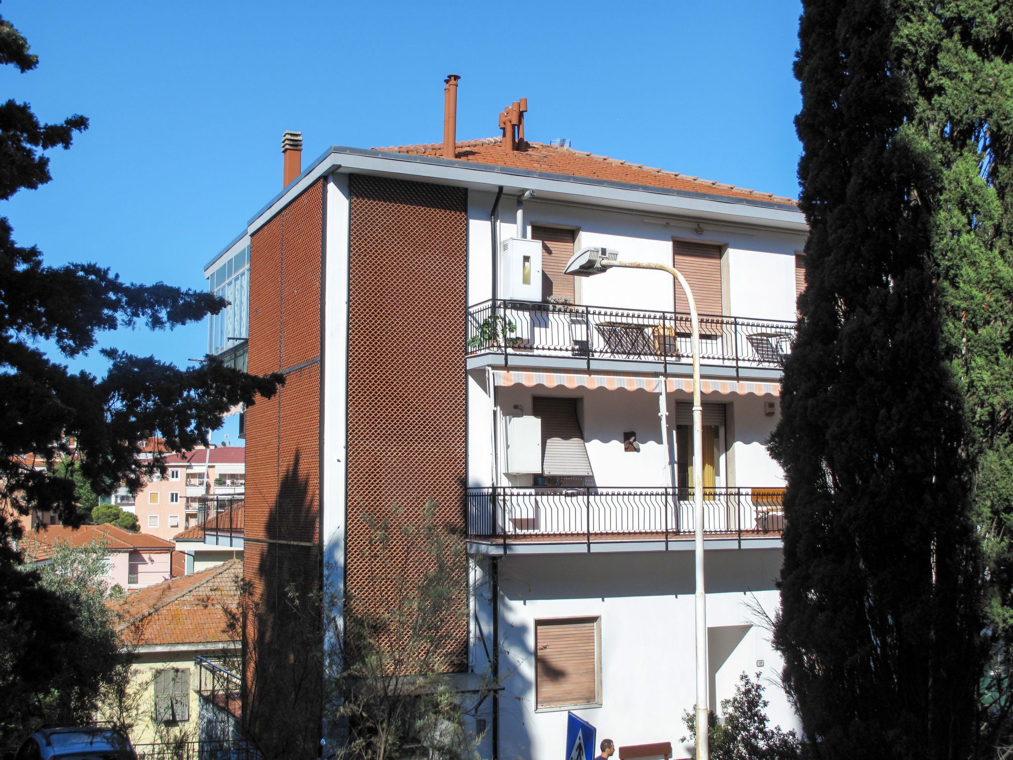 Un edificio de varios pisos con un balcón en el segundo nivel, una chimenea visible y una vista a la calle con árboles y otros edificios al fondo.