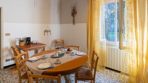 A dining area with a round table set for four, surrounded by wooden chairs. A coffee maker, a bowl of fruit, and a vase with dried flowers are on the table. The room has a tiled floor and a window with yellow curtains. - Cipressi by Interhome (San Lorenzo al Mare)