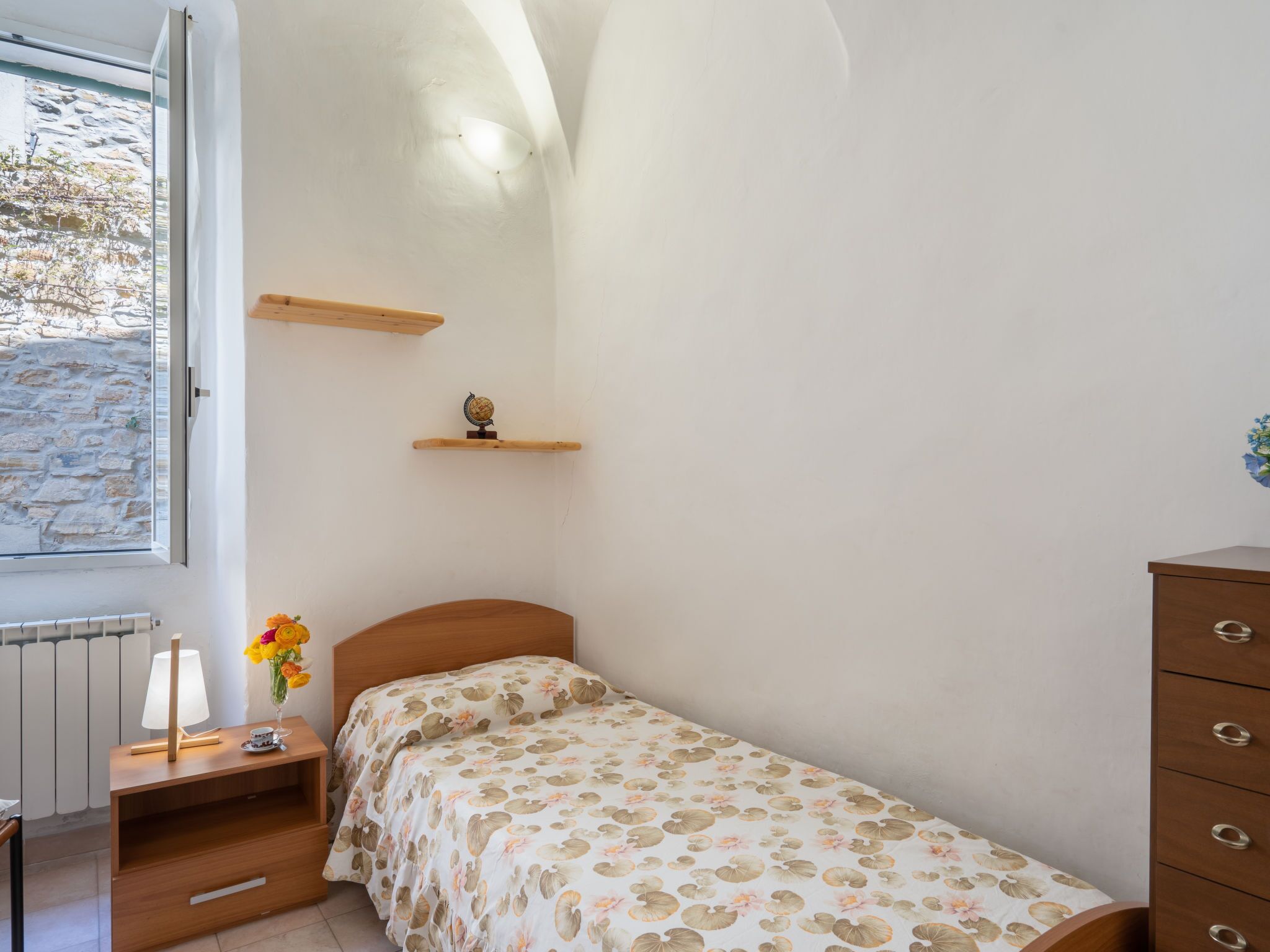 Een kleine, goed verlichte slaapkamer met een enkel bed, een houten nachtkastje, een dressoir, een plank met een wereldbol en een raam met uitzicht op een stenen muur.