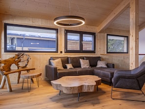 A modern interior with a wooden ceiling, large windows, a grey sectional sofa, a round coffee table, and a unique wooden sculpture. - 02_NTH008_Sup Chalet mit Sauna und Hot Tub 2 Etage by Interhome (Turracher Höhe)
