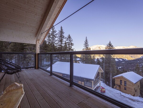 Una terraza de madera con barandilla de vidrio que ofrece vistas a un paisaje nevado de montañas.