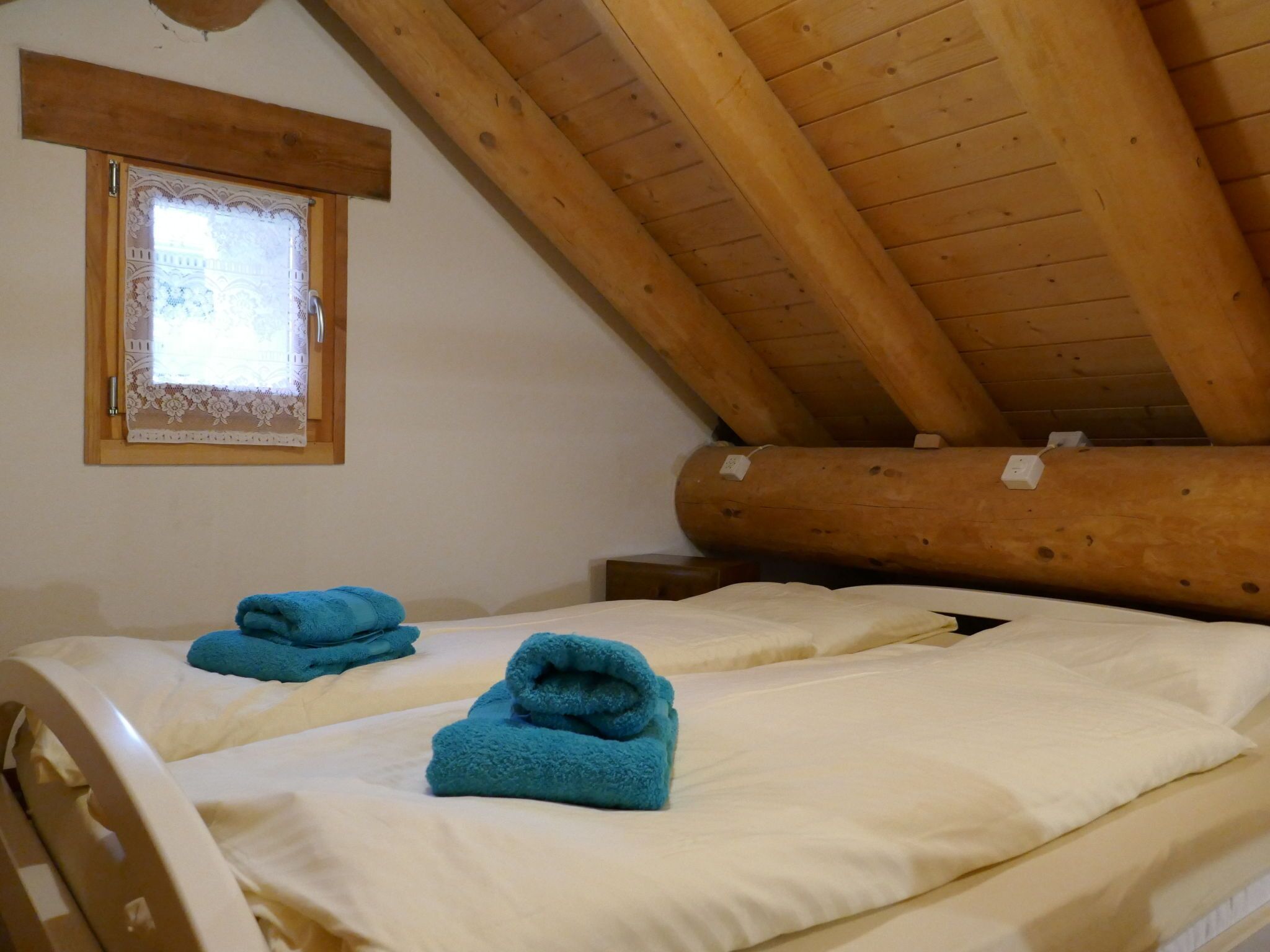 Un dormitorio con dos camas, vigas de madera, una ventana con cortinas de encaje y estructura de troncos de madera.