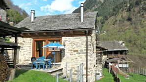 Ein Steinhaus mit Holzschindeln, einem überdachten Außenbereich mit blauen Stühlen und einem Berg im Hintergrund.