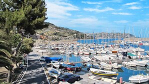 Ein Yachthafen mit zahlreichen vor Anker liegenden Booten, eine Straße mit geparkten Autos und eine hügelige Landschaft im Hintergrund.
