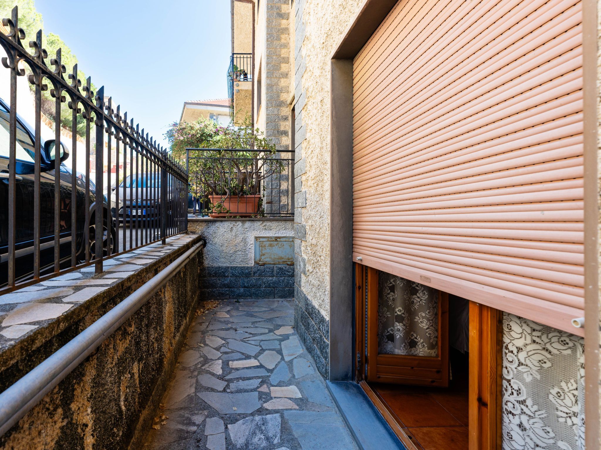 Une ruelle étroite avec un pavé de pierre, une clôture en métal, un pot de fleurs et un bâtiment dont le volet est fermé.