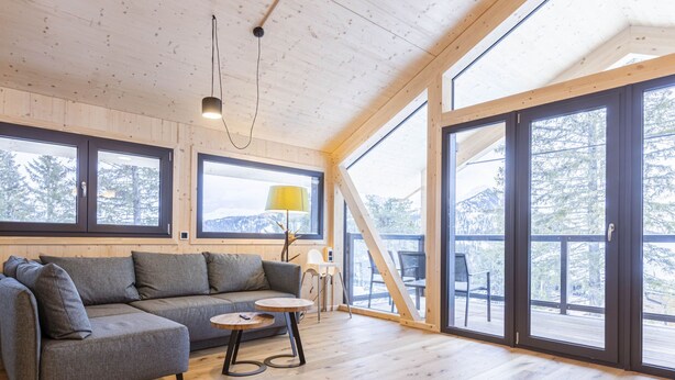 Un intérieur moderne avec un plancher en bois, un canapé gris et de grandes fenêtres offrant une vue sur les arbres et les montagnes.