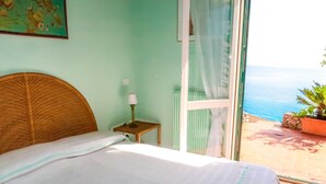 A bedroom with a bed, a nightstand with a lamp, a window with curtains, and a view of the sea. - Paradise View by Interhome (Ventimiglia)
