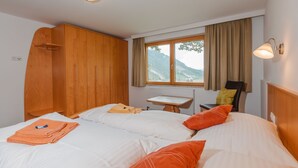 Ein Hotelzimmer mit zwei Betten, einem Holzschrank, einem kleinen Tisch und Blick auf Berge durch ein Fenster.