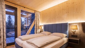 Una cama doble con cabecera, dos mesitas de noche con lámparas, una ventana con cortinas y vistas a árboles.