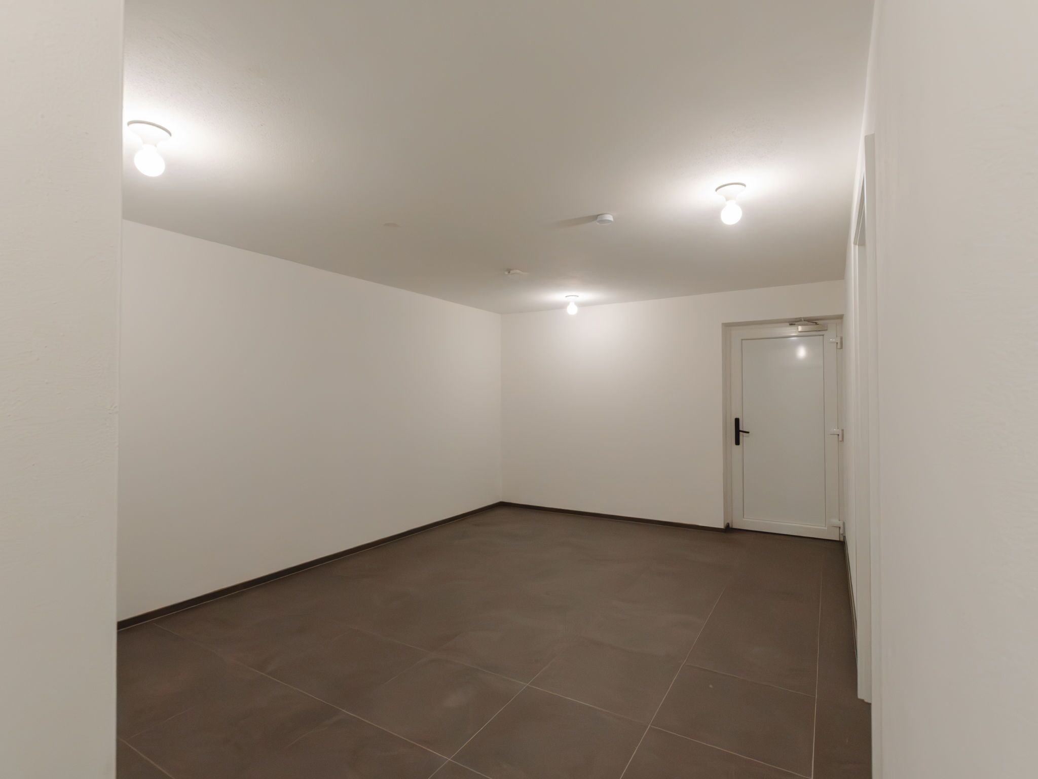A spacious, empty room with white walls and a brown tiled floor. There are three ceiling lights and a door on the right side.