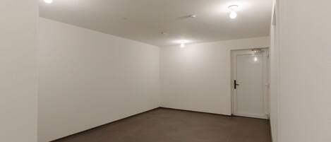 A spacious, empty room with white walls and a brown tiled floor. There are three ceiling lights and a door on the right side.