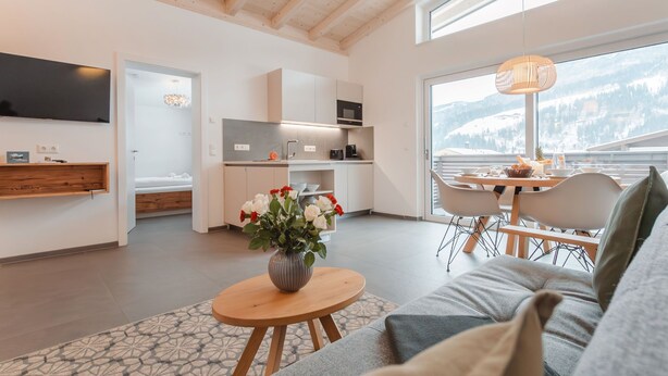 A modern living room with a sofa, a dining table with chairs, a kitchen area, and a large window with a view of a snowy landscape. - Das Georgen, Kitzsteinhorn by Interhome (Bruck)