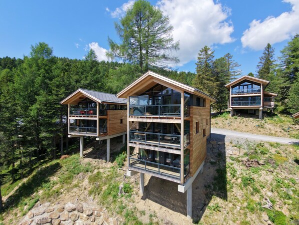 A modern wooden cabin with multiple levels, large windows, and a balcony, surrounded by a forested landscape.