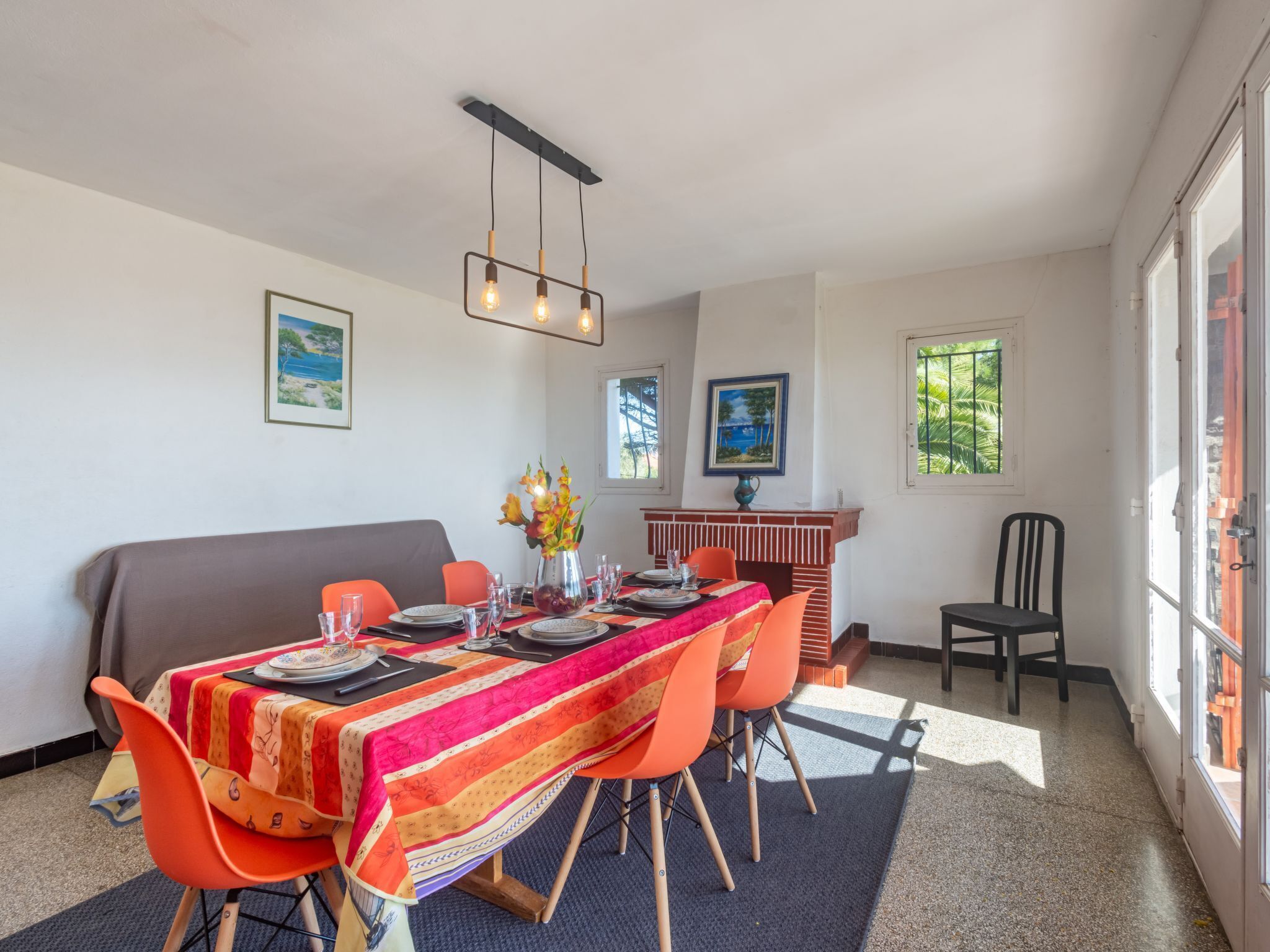 A dining area with a long table, orange chairs, and a colorful tablecloth. There are framed pictures on the walls and a window with a view of greenery.