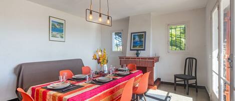 A dining area with a long table, orange chairs, and a colorful tablecloth. There are framed pictures on the walls and a window with a view of greenery.