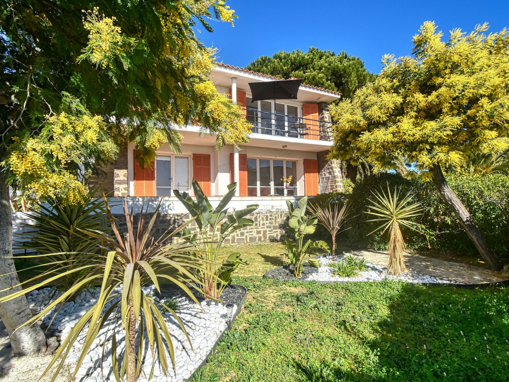 A two-story house with a balcony, surrounded by various plants and trees.