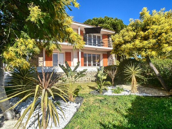 A two-story house with a balcony, surrounded by various plants and trees.