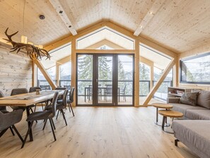 A spacious room with a high ceiling, wooden beams, a large sliding glass door leading to a balcony, and a dining area with a table and chairs.