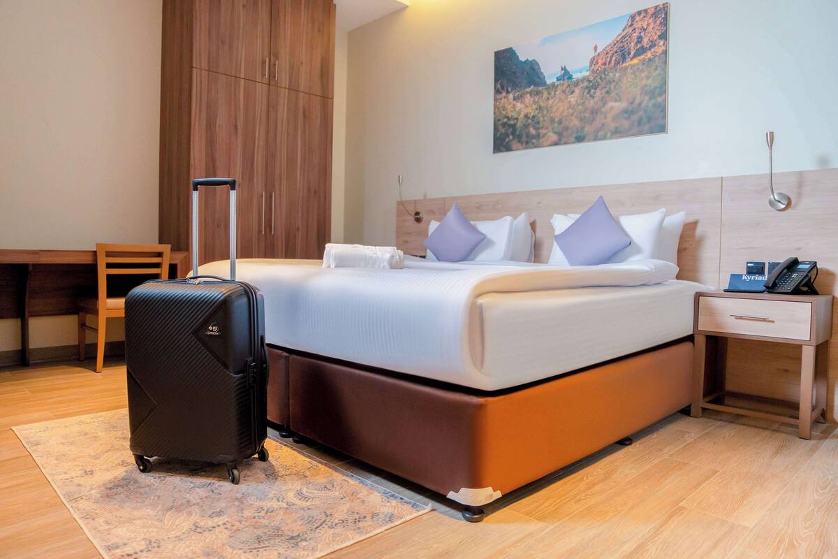 A hotel room with a bed, a suitcase, a desk, and a nightstand with a phone.