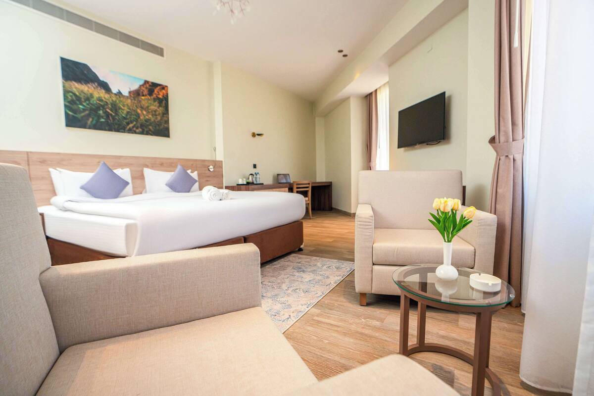 A hotel room with a bed, a sofa, a small table with a vase of flowers, a TV, and a painting on the wall.