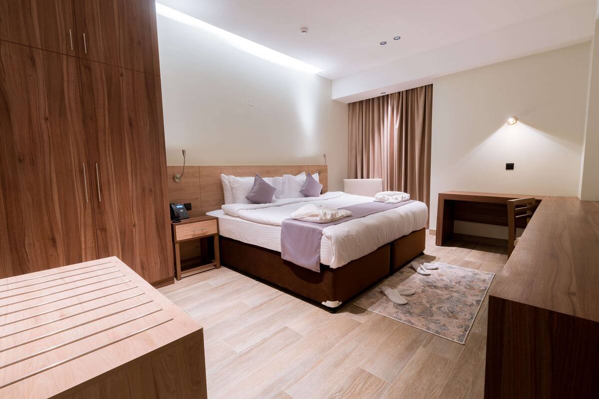 A modern hotel room with a wooden bed, a bedside table, a desk, and a wardrobe.