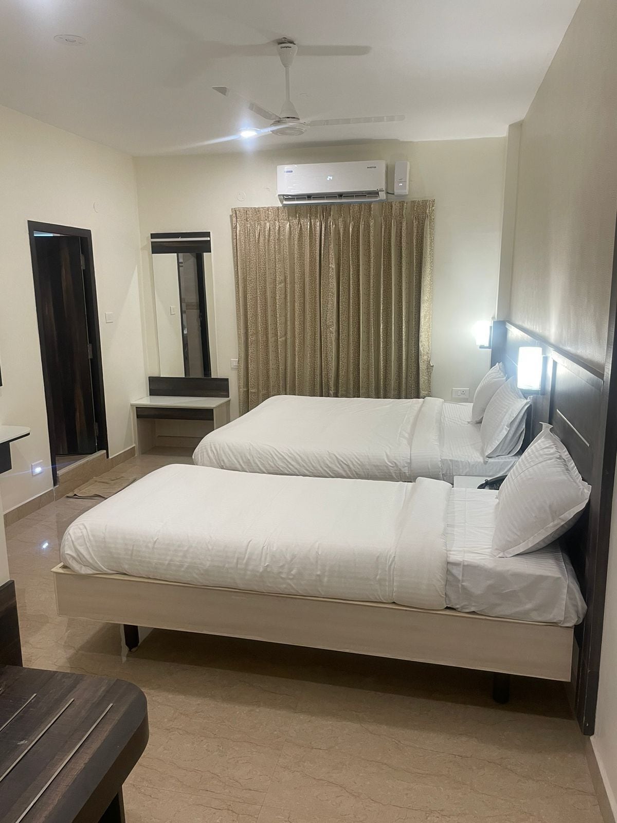 A hotel room with a bed, a desk, a chair, a mirror, a ceiling fan, and an air conditioning unit.