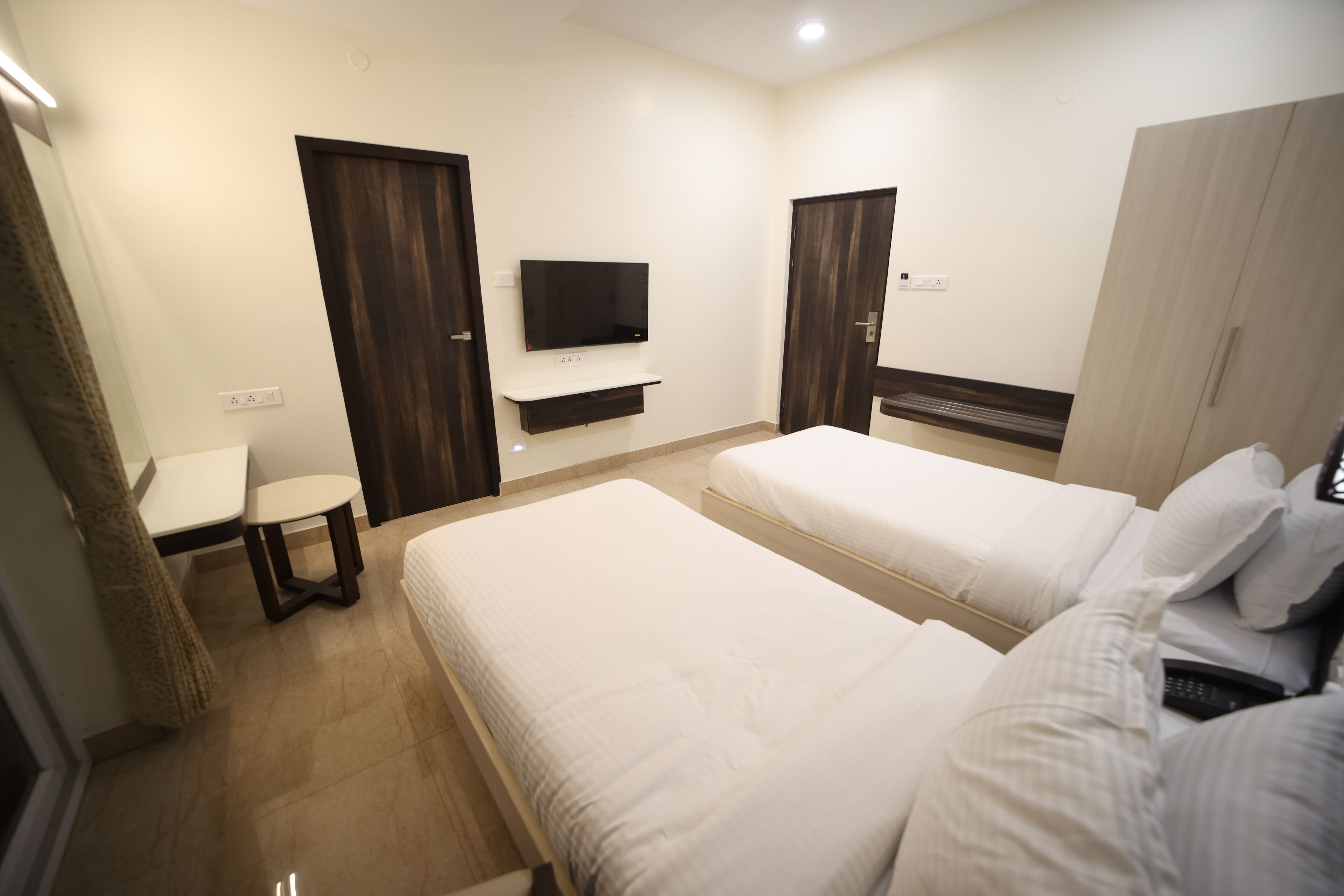A hotel room with two beds, a TV mounted on the wall, a small table, and a wardrobe.