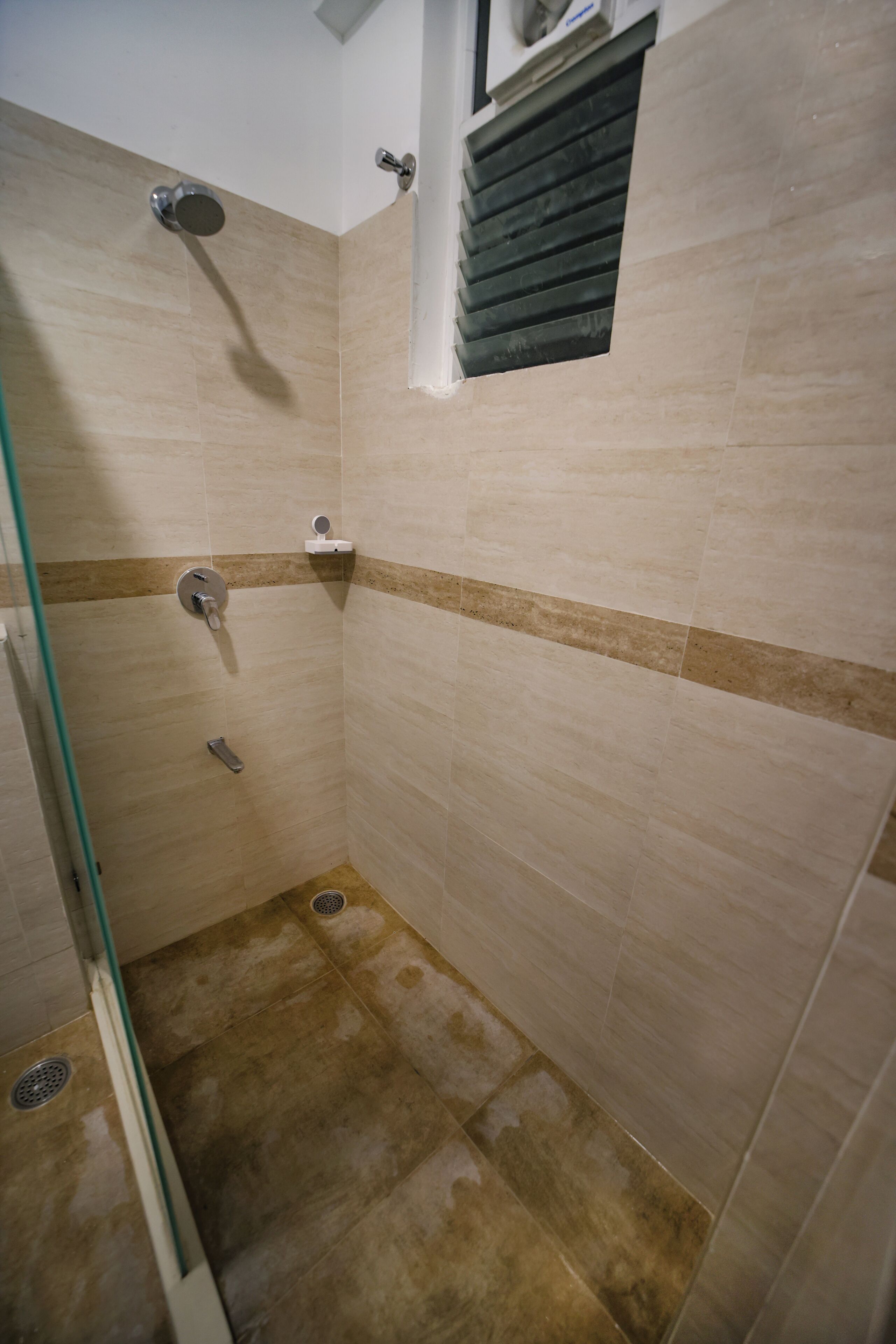 Un petit espace de douche carrelé, doté d’une fenêtre et d’une grille de ventilation.